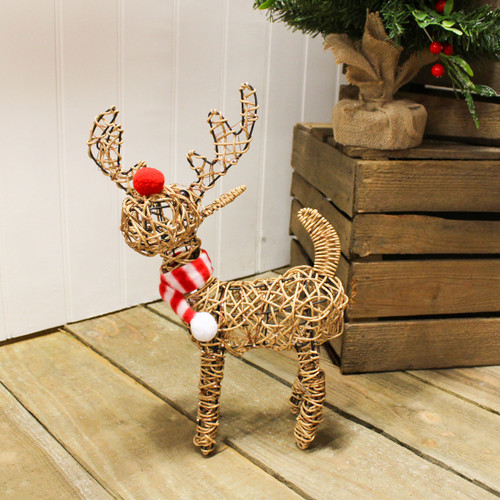 Wicker reindeer Christmas decoration with red nose and striped scarf, standing on a wooden floor beside a rustic wooden crate and a small artificial tree with red berries.