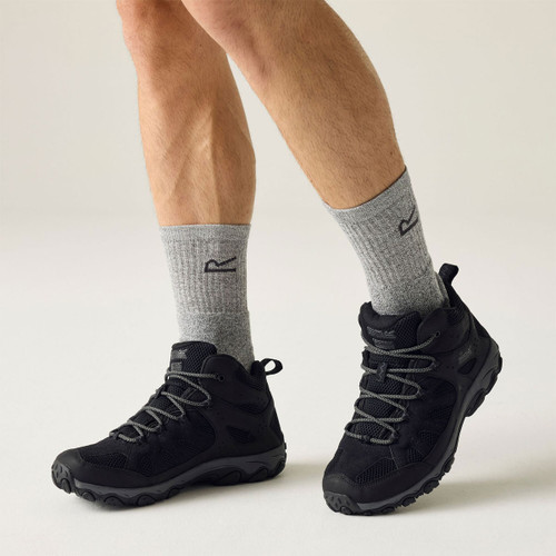 Black mid-height walking shoes with grey laces and rugged soles, worn with grey hiking socks in a studio setting.