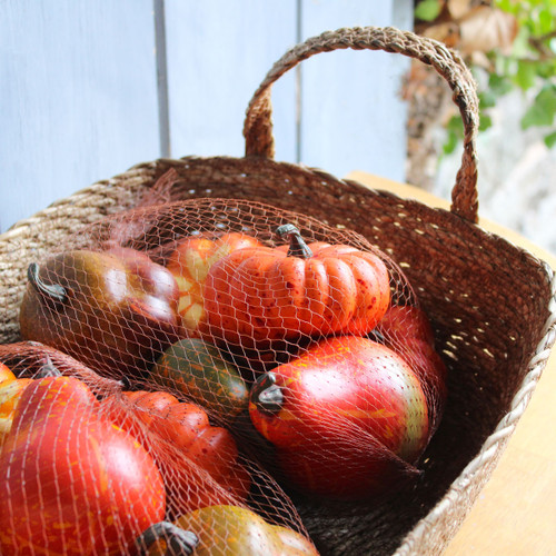 A woven basket filled with artificial pumpkins and gourds in orange, red, and green tones, wrapped in net packaging.