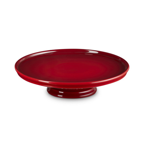 A Le Creuset stoneware cake stand in Cerise red, shown at an angle highlighting the wide glossy surface and sturdy base.