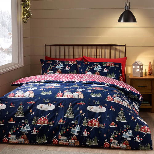 A cosy bedroom with Christmas bedding featuring a navy duvet cover decorated with festive village scenes, including snowy houses, Christmas trees, ice skaters, and snowmen, with a red and white check reverse.