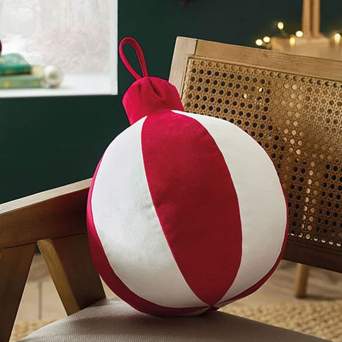 A red and white bauble-shaped cushion with a fabric loop on top, styled on a wooden chair with festive decor in the background.