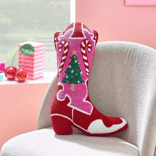 A decorative cushion shaped like a cowboy boot in festive colours, featuring a Christmas tree and candy cane design in pink, red, green, and white. Displayed on a light grey chair in a cheerful room.