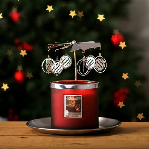 Red Christmas candle with silver carousel topper decorated with hanging bauble ornaments, glowing warmly in front of a blurred Christmas tree with gold star-shaped lights.