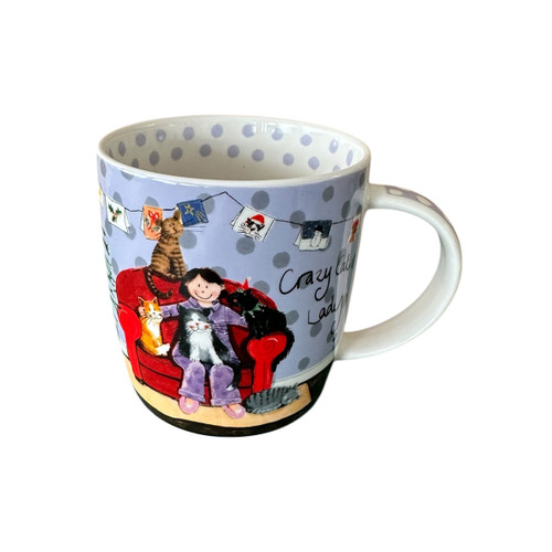 White mug with a lilac background and grey polka dots, featuring a cartoon of a smiling woman in purple pyjamas on a red armchair surrounded by cats. Above her is a string of festive cards, with the phrase “Crazy Cat Lady” written to the side. The inside rim is decorated with grey spots.