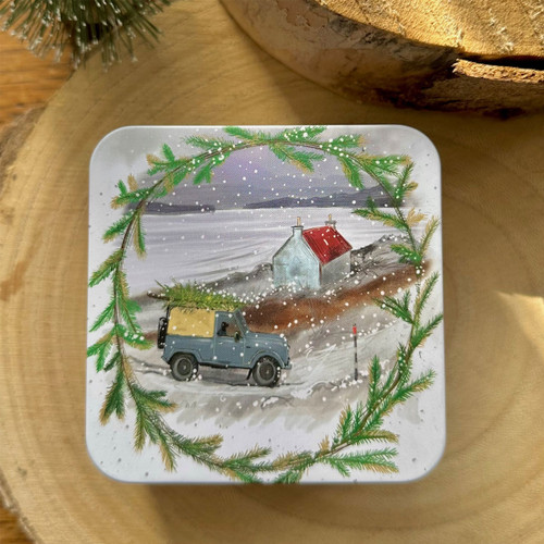 Square tin decorated with a snowy countryside scene featuring a blue Land Rover carrying a Christmas tree, with a cottage in the background, framed by green fir branches.