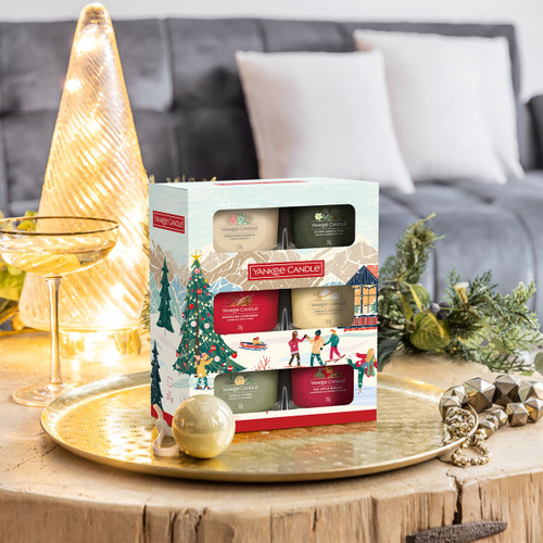 A Yankee Candle votive gift set displayed on a golden tray with Christmas decorations. The six-piece candle selection is presented in a festive illustrated box, placed in a cosy living room with seasonal décor.