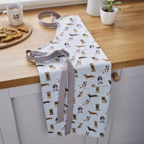 Curious Dogs cotton apron with illustrated dog print, displayed on a kitchen counter with biscuits and a mug.