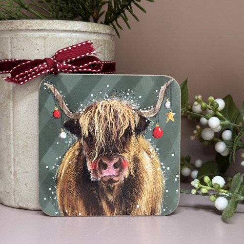 A festive square coaster featuring an illustrated Highland cow with long shaggy hair, decorated with Christmas baubles and a gold star hanging from its horns, set against a green background with falling snow.