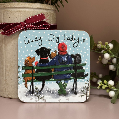 A square coaster featuring an illustration of a woman in a red bobble hat and blue coat sitting on a green bench with four dogs, watching the snowfall. The text above reads “Crazy Dog Lady” in a handwritten style.