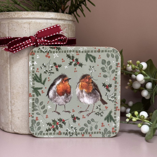 A square coaster decorated with an illustration of two robins facing each other. The birds have red breasts and fluffy feathers, set against a sage green background patterned with holly, berries, fir branches and white stars for a festive design.