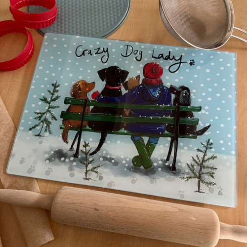 Rectangular glass worktop saver featuring an illustration of a woman in a red bobble hat and green wellies sitting on a green bench with four dogs in the snow, captioned "Crazy Dog Lady".