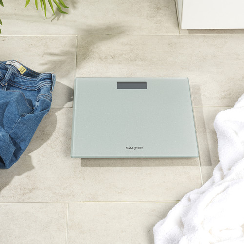 Digital bathroom scale on beige tiles, flanked by folded jeans on the left and a white towel on the right, conveying a sense of daily routine.
