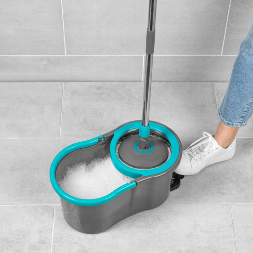 A person in jeans and white sneakers, stepping on a grey and blue spin mop bucket on a tiled floor, creating soapy foam in the bucket.