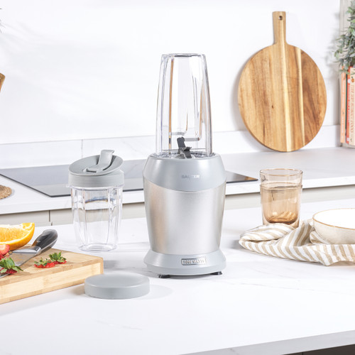 A silver blender sits on a white worktop beside a transparent travel cup and a glass. A wooden cutting board with vegetables is in the background.