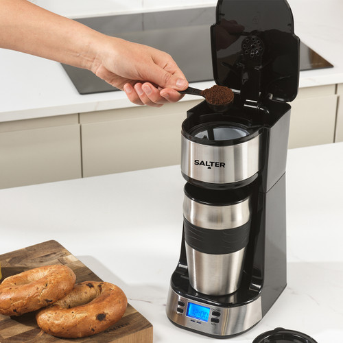 A hand pours ground coffee into a Salter coffee maker on a kitchen counter. Nearby, two bagels rest on a cutting board. The scene feels inviting.