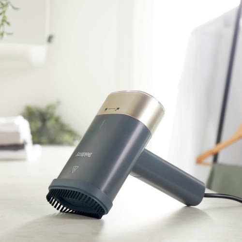 A sleek, modern lint remover with a matte grey body and a gold accent is placed on a white surface. The background is softly blurred with a white shirt on a hanger, conveying a tidy and minimalist aesthetic.