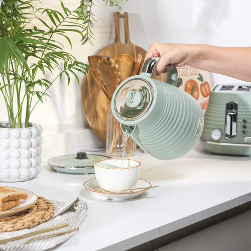 A hand pours water from a stylish mint green kettle into a white cup on a kitchen counter. Nearby, there's a matching toaster, lush plant, and wooden utensils, creating a cosy, fresh atmosphere.