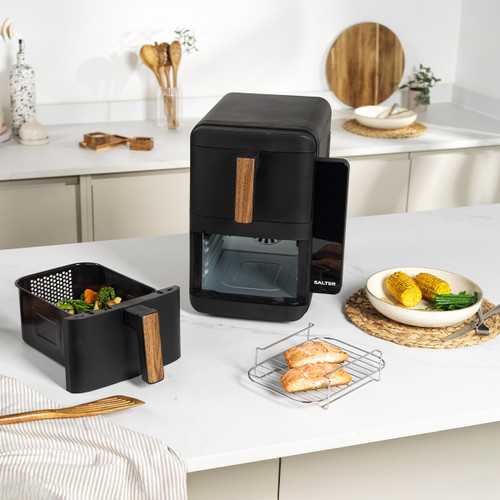 A sleek black air fryer with wood accents sits on a white kitchen counter. Nearby, a basket with vegetables, a rack with chicken, and a plate with corn and beans.