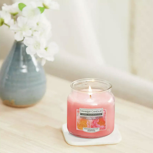 Yankee Candle Home Inspiration Coral Peony scented jar candle burning on a white coaster beside a vase of white flowers.