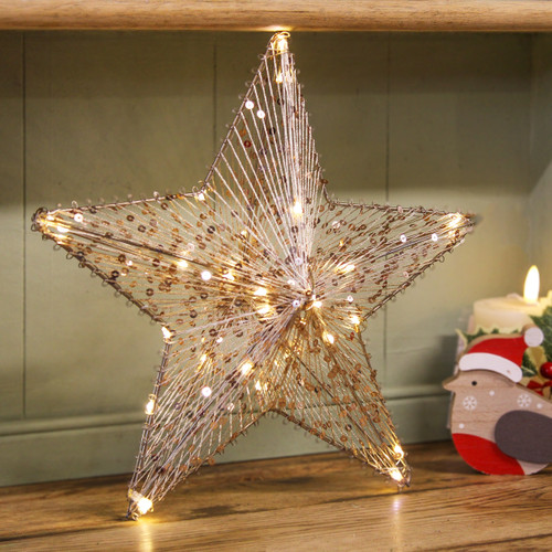 A five-pointed Christmas star decoration made from silver thread and gold sequins, illuminated by warm white fairy lights, standing on a wooden shelf beside a festive candle.