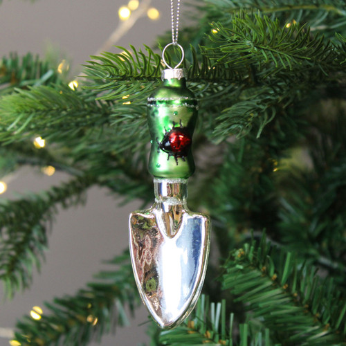 A silver and green Christmas tree decoration shaped like a small garden trowel, hanging from a green fir branch.
