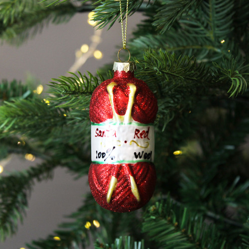 A sparkling red yarn ball Christmas decoration, shaped like a ball of wool, hanging from a green Christmas tree branch with warm fairy lights in the background.