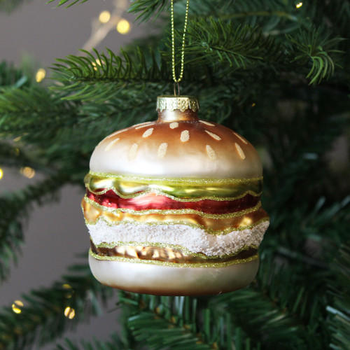 A glass Christmas ornament shaped like a burger, with a golden bun, glittery sesame seeds. It hangs from a green Christmas tree branch with soft fairy lights in the background.