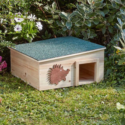A small wooden hedgehog house sits on green grass, surrounded by lush plants and flowers. The house features a cut-out hedgehog design on its side.