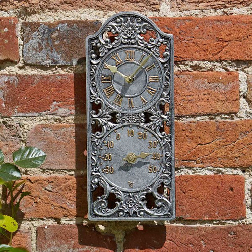 Ornate wall-mounted clock and thermometer on a red brick wall. The clock features intricate metalwork and displays Roman numerals, conveying a vintage charm.