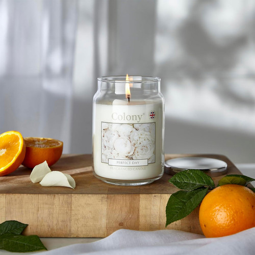 Colony Perfect Day fragranced candle lit on a wooden surface beside fresh oranges and petals, creating a bright, calming home atmosphere with citrus and floral accents.