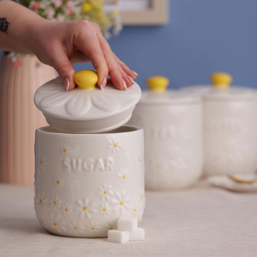 Hand lifting the daisy-patterned ceramic lid from the Price & Kensington sugar jar with sugar cubes on the table and matching jars in the background.