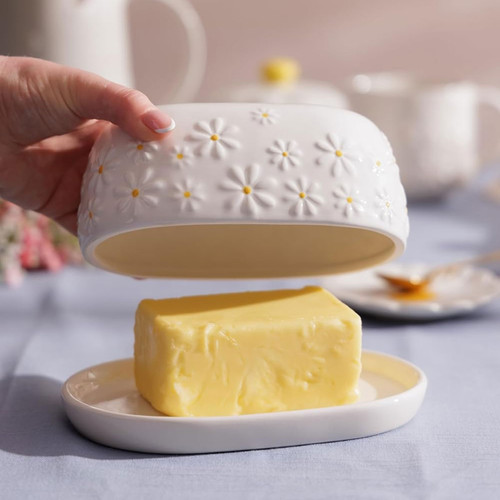 Hand lifting the daisy-patterned ceramic lid from the Price & Kensington butter dish, revealing a block of butter on the matching tray.