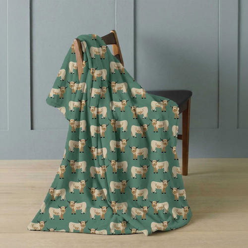 Green Highland cow print throw blanket draped over a wooden chair, featuring a repeating cream and brown Highland cow design.