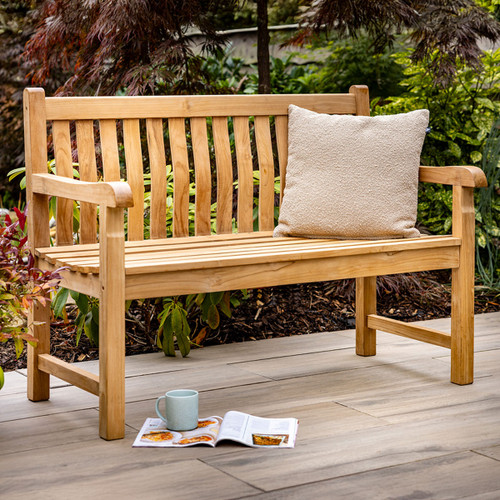 Wooden outdoor garden bench made from natural teak, shown with a textured beige cushion on the seat and surrounded by lush green plants.