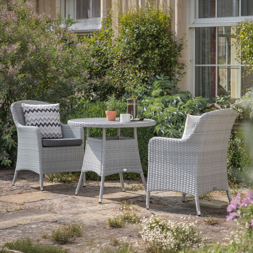 Outdoor bistro set with two grey wicker armchairs and a round matching table placed on a stone patio, surrounded by lush garden plants.
