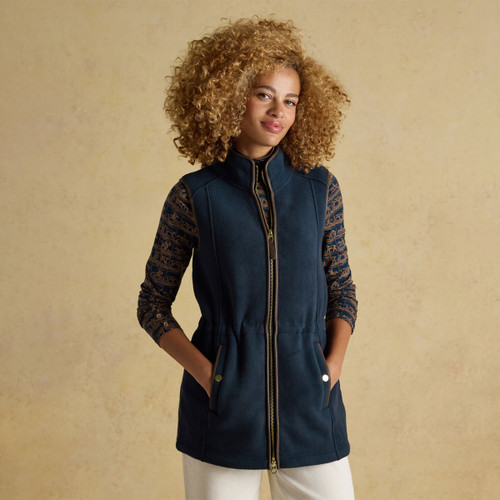 A women’s longline navy fleece gilet with a high collar, full-length zip and adjustable waist, styled with a printed top and white trousers.