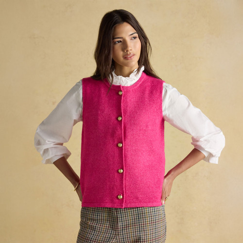 Model wearing a bright pink sleeveless knitted vest with gold textured buttons, styled over a white ruffled blouse and checked trousers.