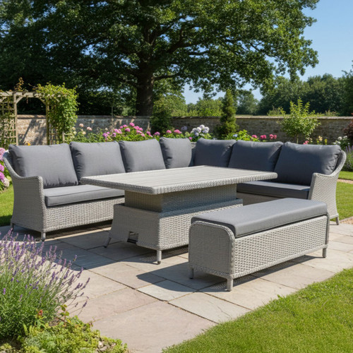 A grey rattan corner garden dining set with deep grey cushions, a rectangular dining table and a matching cushioned bench, arranged on a stone patio surrounded by colourful flowers and greenery.