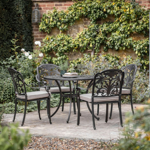 Bronze 4-seat garden dining set on a stone patio with a round metal table and four armchairs with light seat cushions, set against a leafy brick wall.