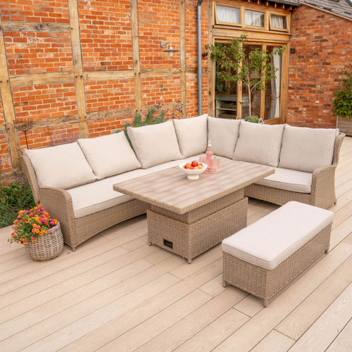 Outdoor corner sofa set with beige cushions and an adjustable rattan table on a light timber deck beside a red-brick wall.