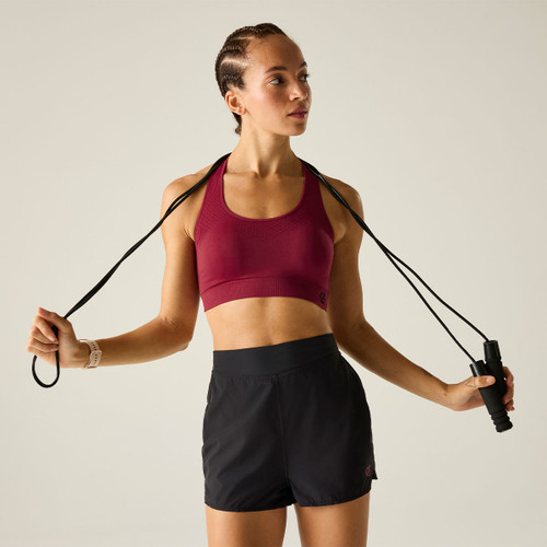 Model wearing a burgundy racerback sports bra and black running shorts, holding a skipping rope in a studio setting.