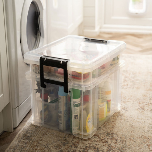 Transparent storage box with black clip handles placed beside a washing machine, filled with assorted household cleaning products.