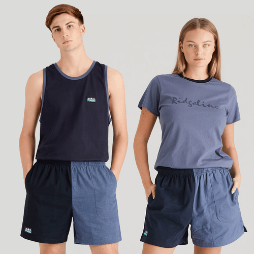 Two models wear matching color-block shorts split into dark navy and light blue halves, with an elastic waistband, side pocket, and small logo on the darker panel.