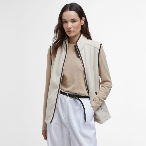 Model wearing a cream Barbour fleece gilet with dark brown trim over a beige jumper and white trousers.