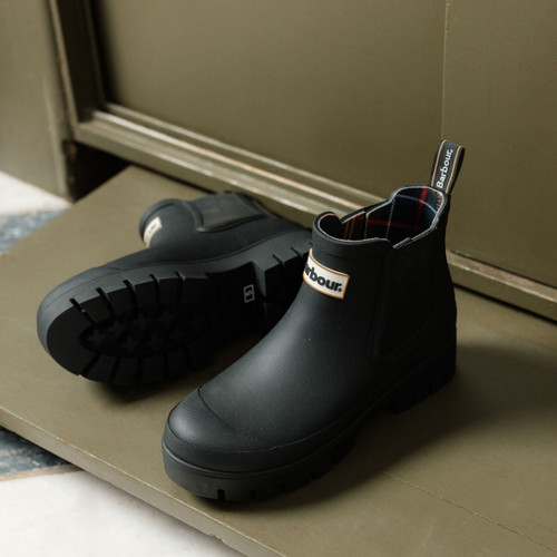Black Barbour Chelsea wellington boots with chunky lug sole, shown on a doorstep with a matte rubber finish and pull tab at the heel.