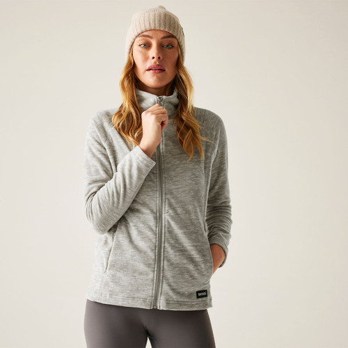 Model wearing a light grey full-zip fleece jacket with a high collar and a cream beanie, holding the zip, against a light background.