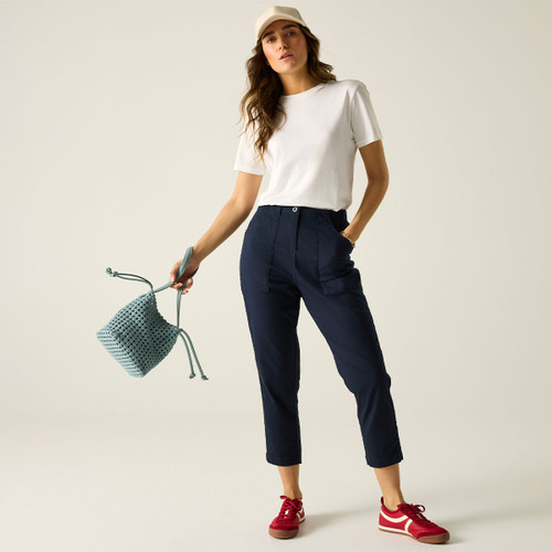 Full-length view of a model wearing navy cropped trousers with a white T-shirt, beige cap and red trainers, holding a small blue bag.