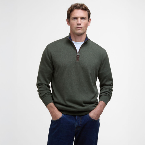 Man wearing a green half-zip jumper over a white shirt, with hands in the pockets of blue jeans. He stands against a plain white background.