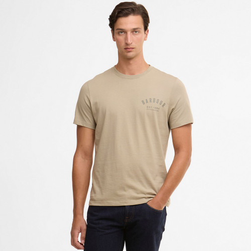 A person in a beige Barbour T-shirt and dark jeans stands against a plain white background. The expression is neutral, conveying a calm, casual tone.